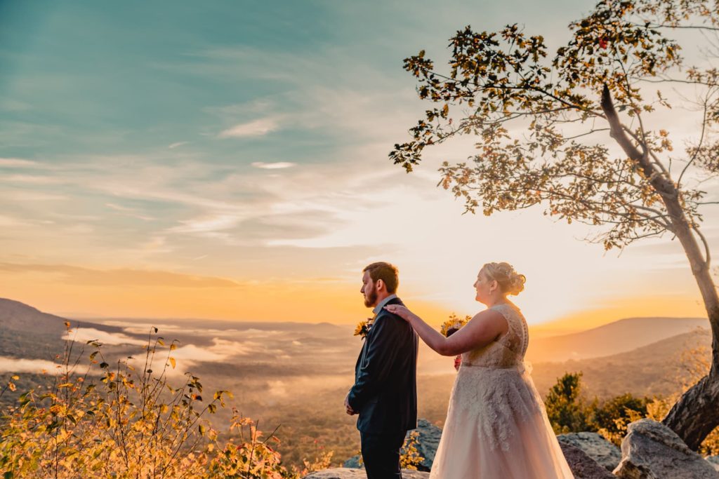The bride, holding her bouquet in one hand, touches her groom on the should as they begin their sunrise first look at this overlook during their Pennsylvania elopement.