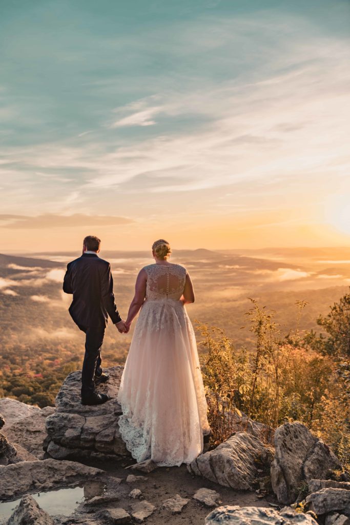 A couple holds hands as they stand on an overlook facing a fog covered valley and a beautiful sunrise.