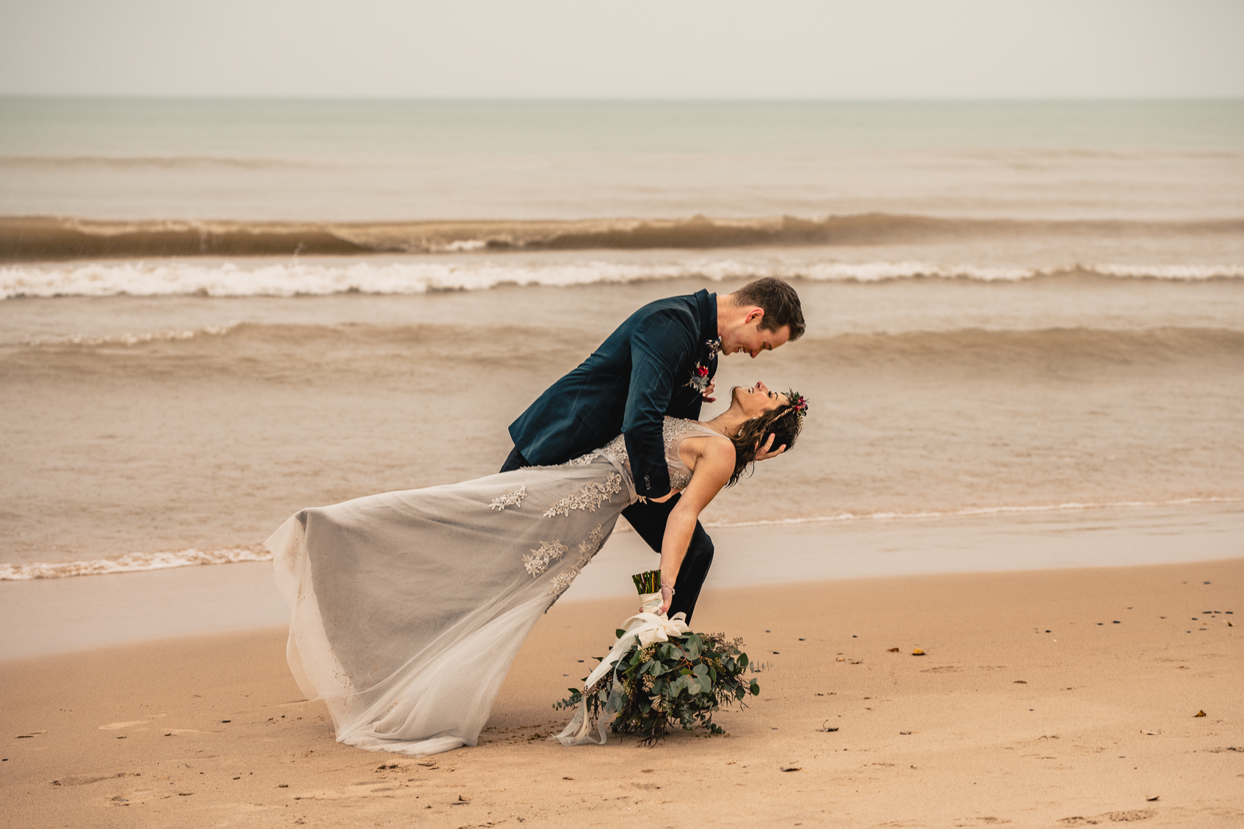 The groom dips the bride as the waves roll in along the beach in this Wisconsin elopement