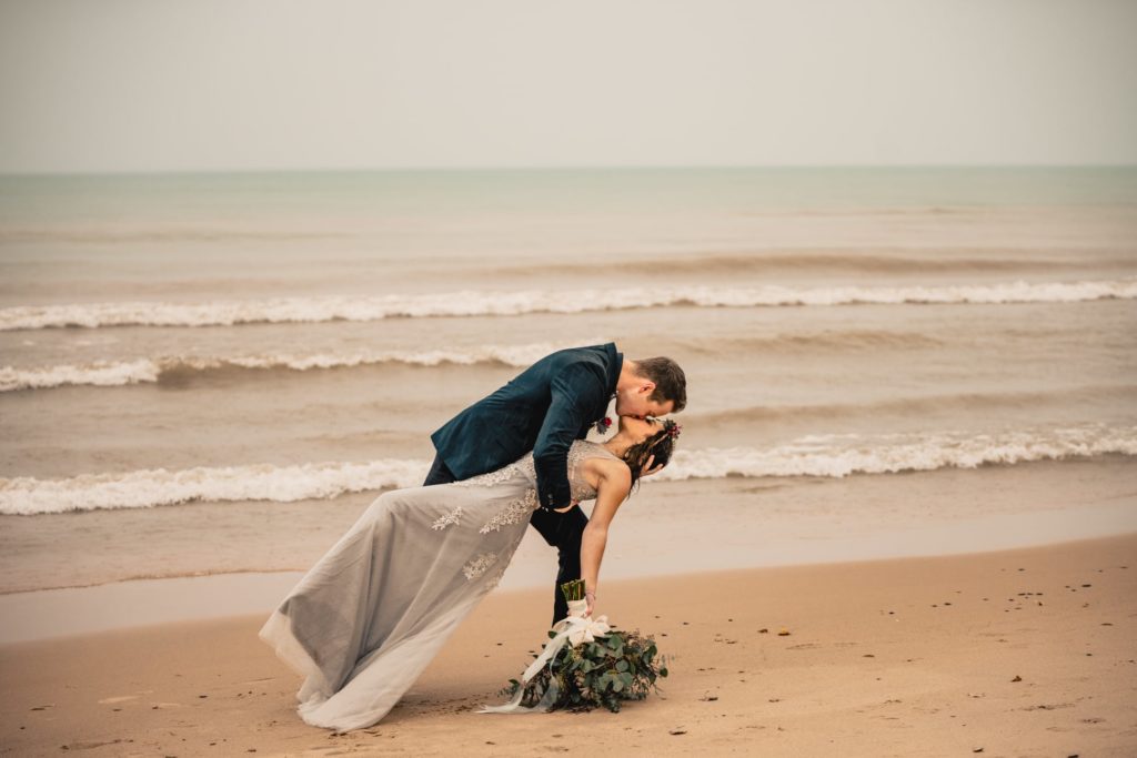 A groom dips his bride in the rain on as the waves roll in on the beach behind them