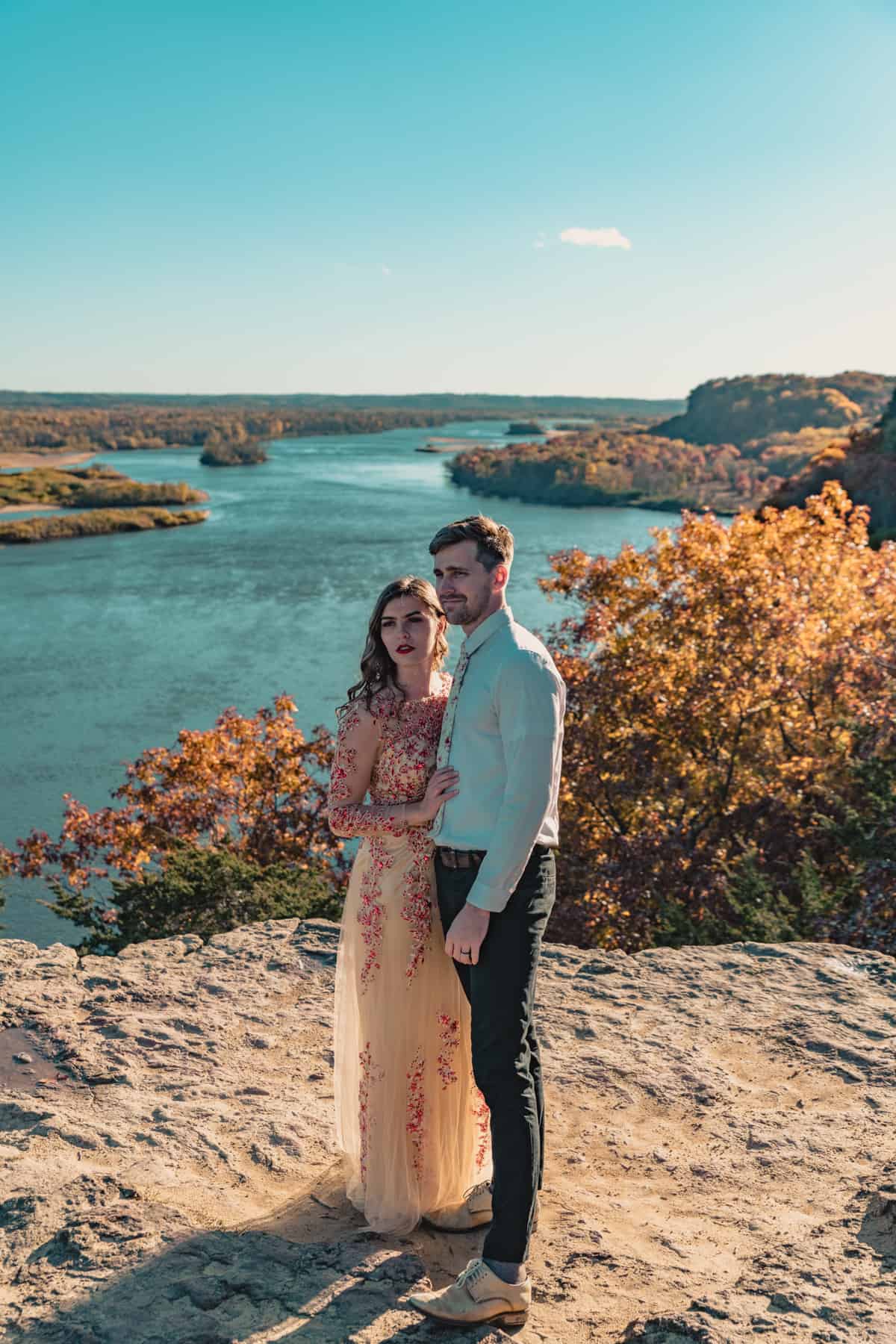 A couple poses a top a cliff with fall colors surrounding them, her in a gold wedding gown, overlooking a flowing river in Wisconsin