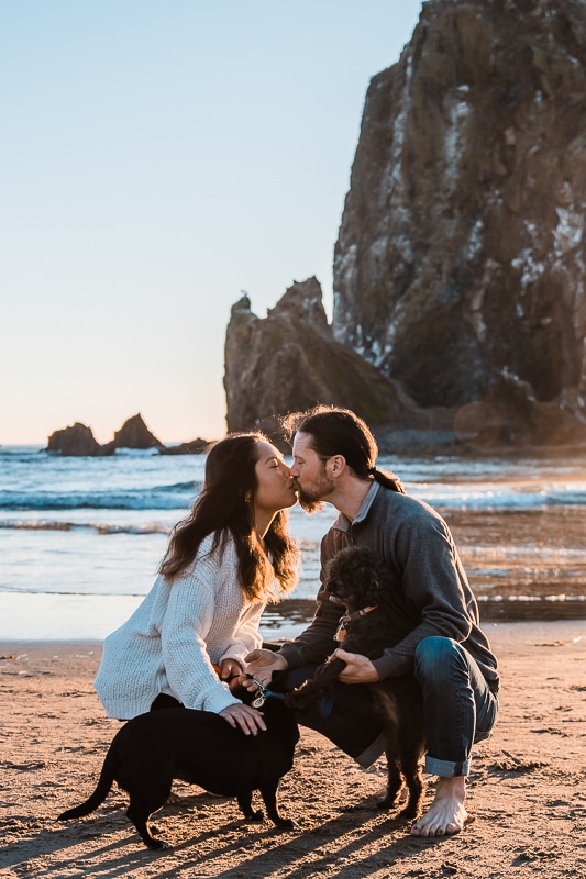 With their dogs in hand for their Oregon Coast engagement session, an engaged couples kisses in front of Haystack Rock on Cannon Beach 