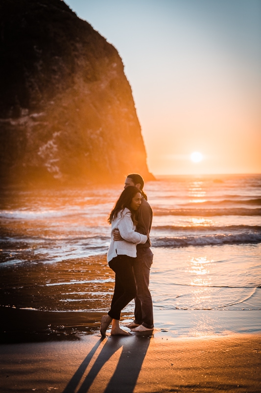 Hugging each other at sunset, this engaged couple poses for their engagement session with Haystack Rock in the background on Cannon Beach on the Oregon Coast