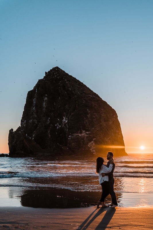 An engaged couple embraces in front of Haystack Rock on Cannon Beach on the Oregon Coast