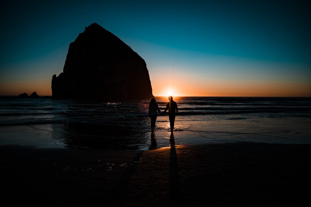 Backlit by the setting sun, an engaged couple walks along Cannon Beach during their Oregon Coast engagement session