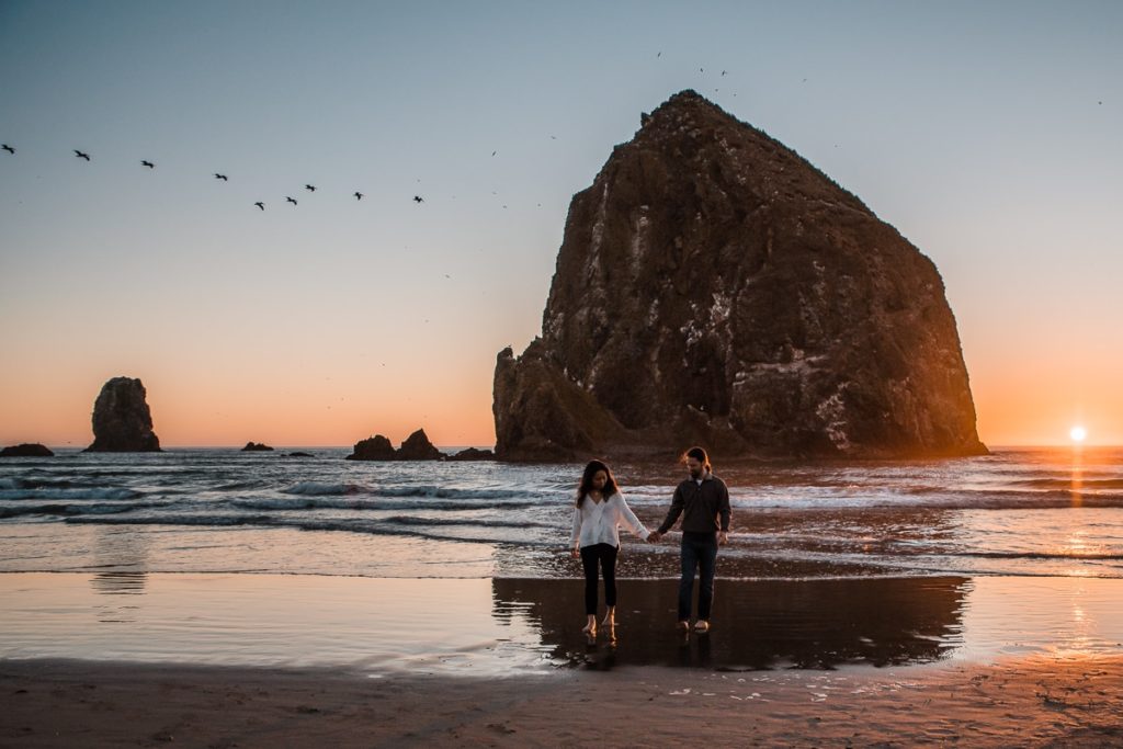With birds flying overhead, an engaged couples walks along Cannon Beach in front of Haystack Rock for their Oregon Coast engagement session