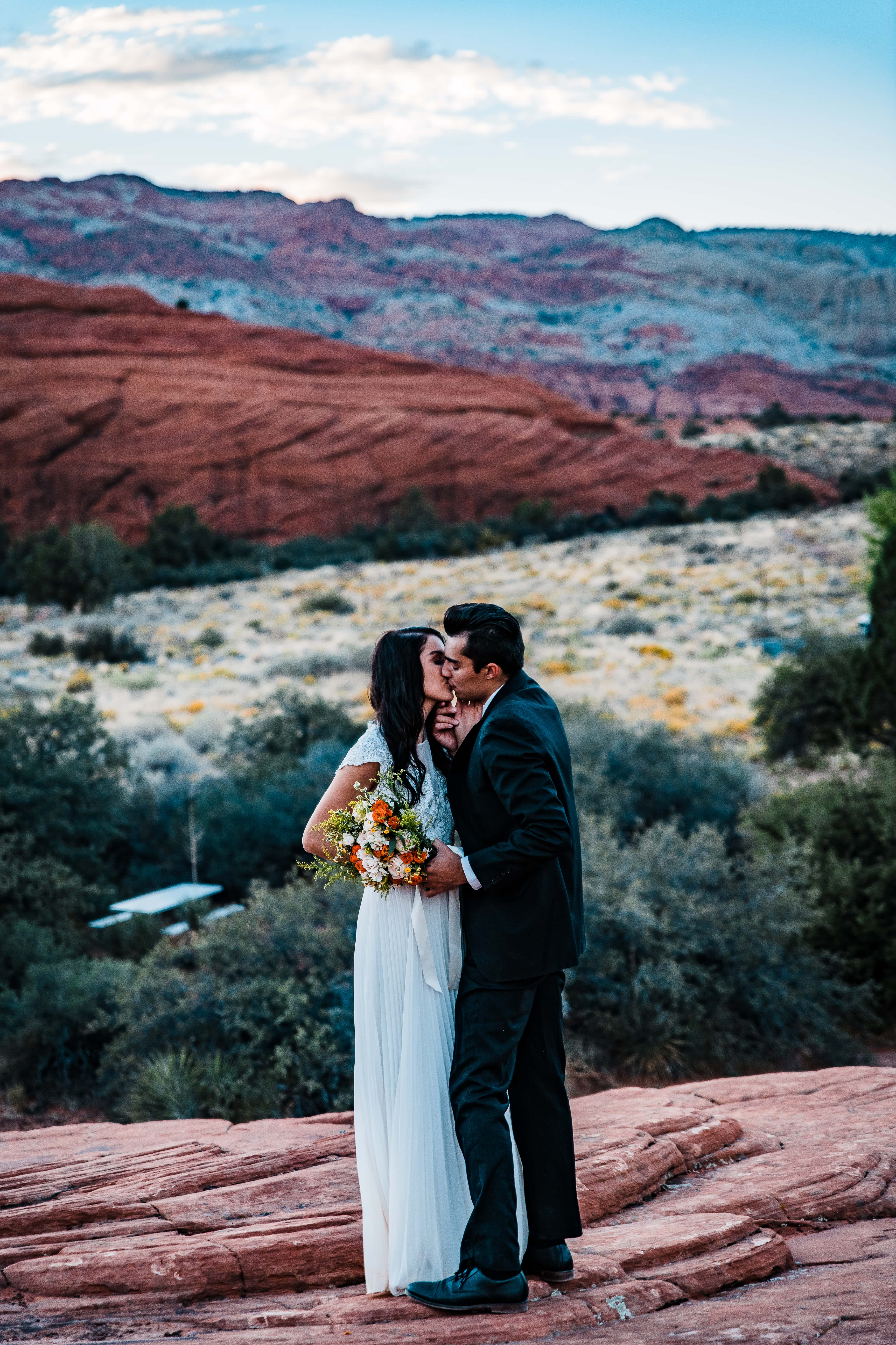 A couple kisses for their wedding portraits in the red rocks of southern Utah, her in a white wedding dress, and him wearing a black suit.